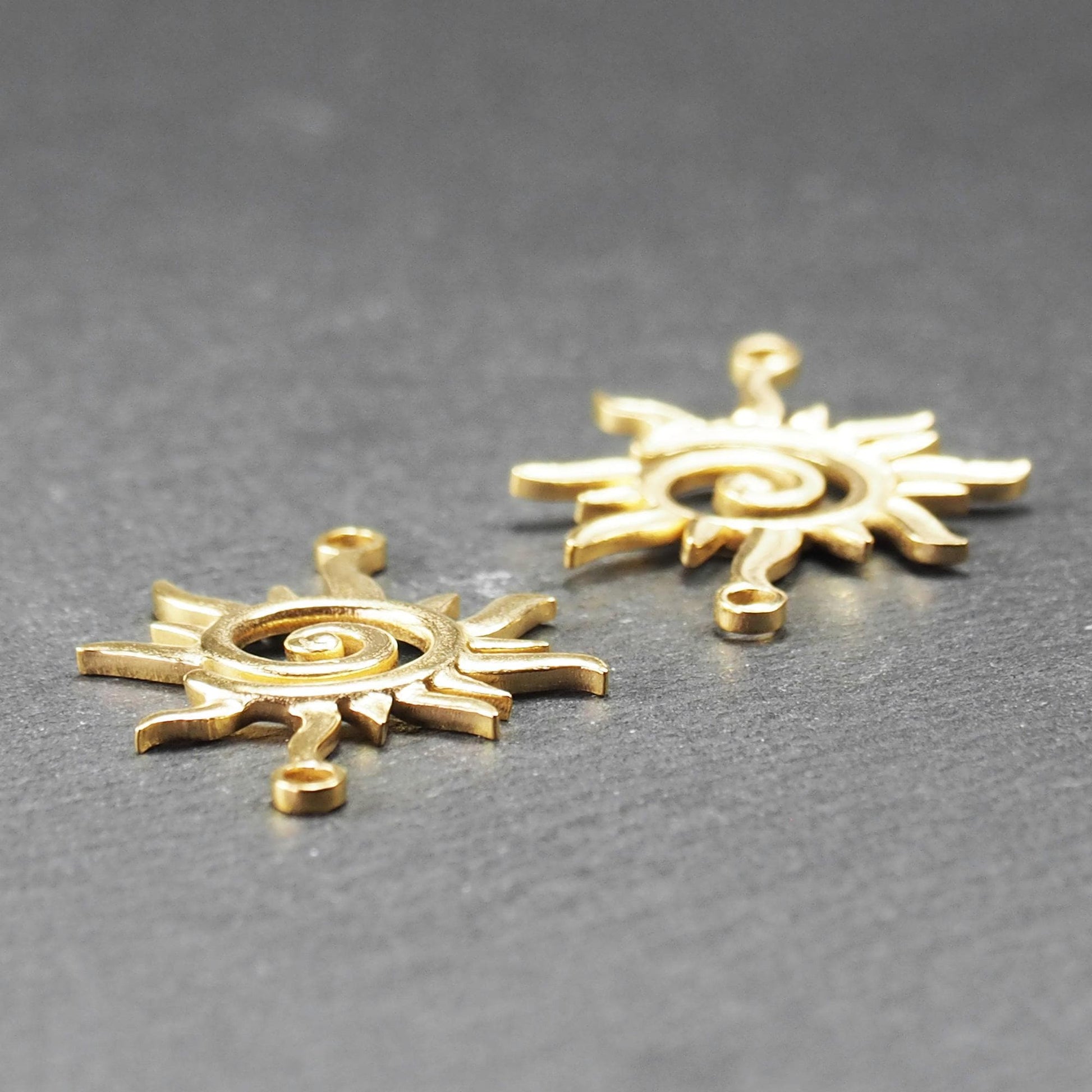 Goldfarbener, durchbrochener Sonnen-Verbinder mit spiralförmigem Zentrum, unregelmäßigen wellenförmigen Strahlen und je einer Öse oben und unten.  Vorder- und Rückseite.