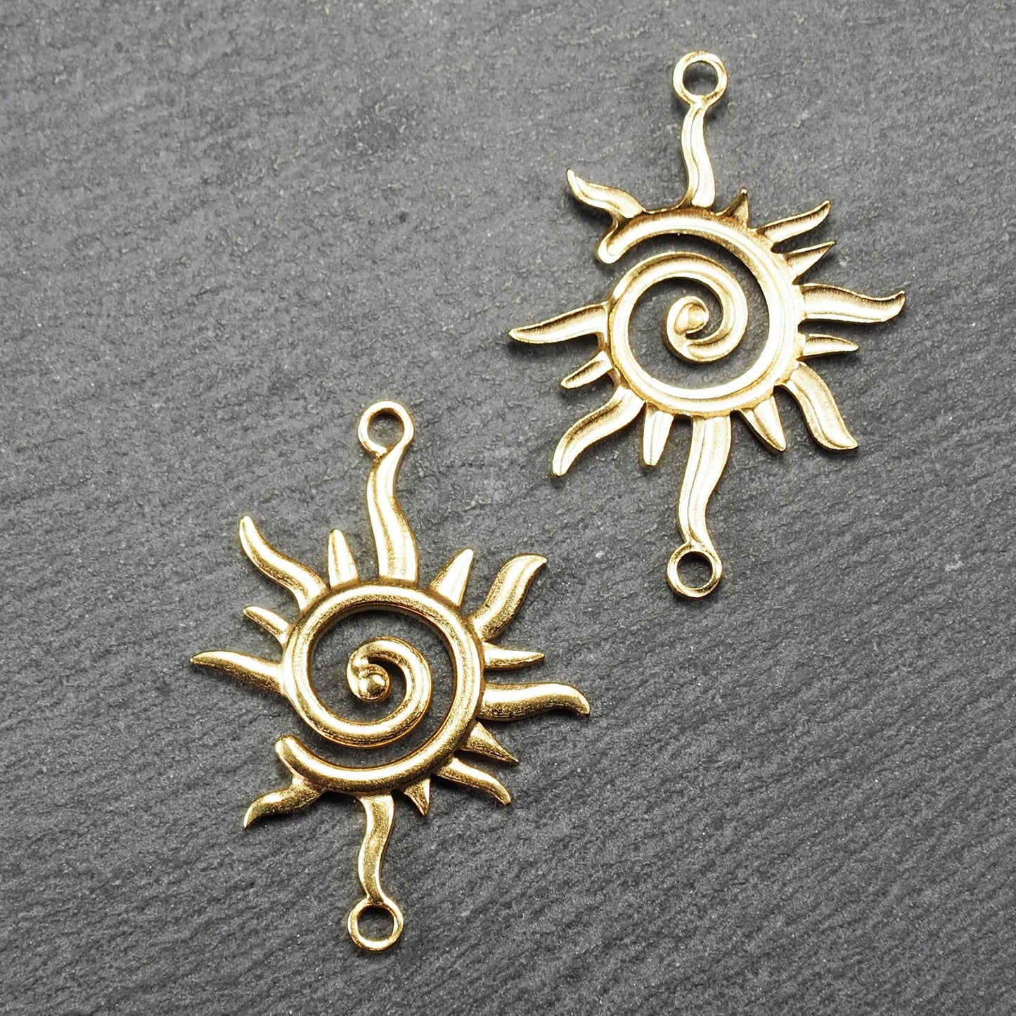 Goldfarbener, durchbrochener Sonnen-Verbinder mit spiralförmigem Zentrum, unregelmäßigen wellenförmigen Strahlen und je einer Öse oben und unten.  Vorder- und Rückseite.