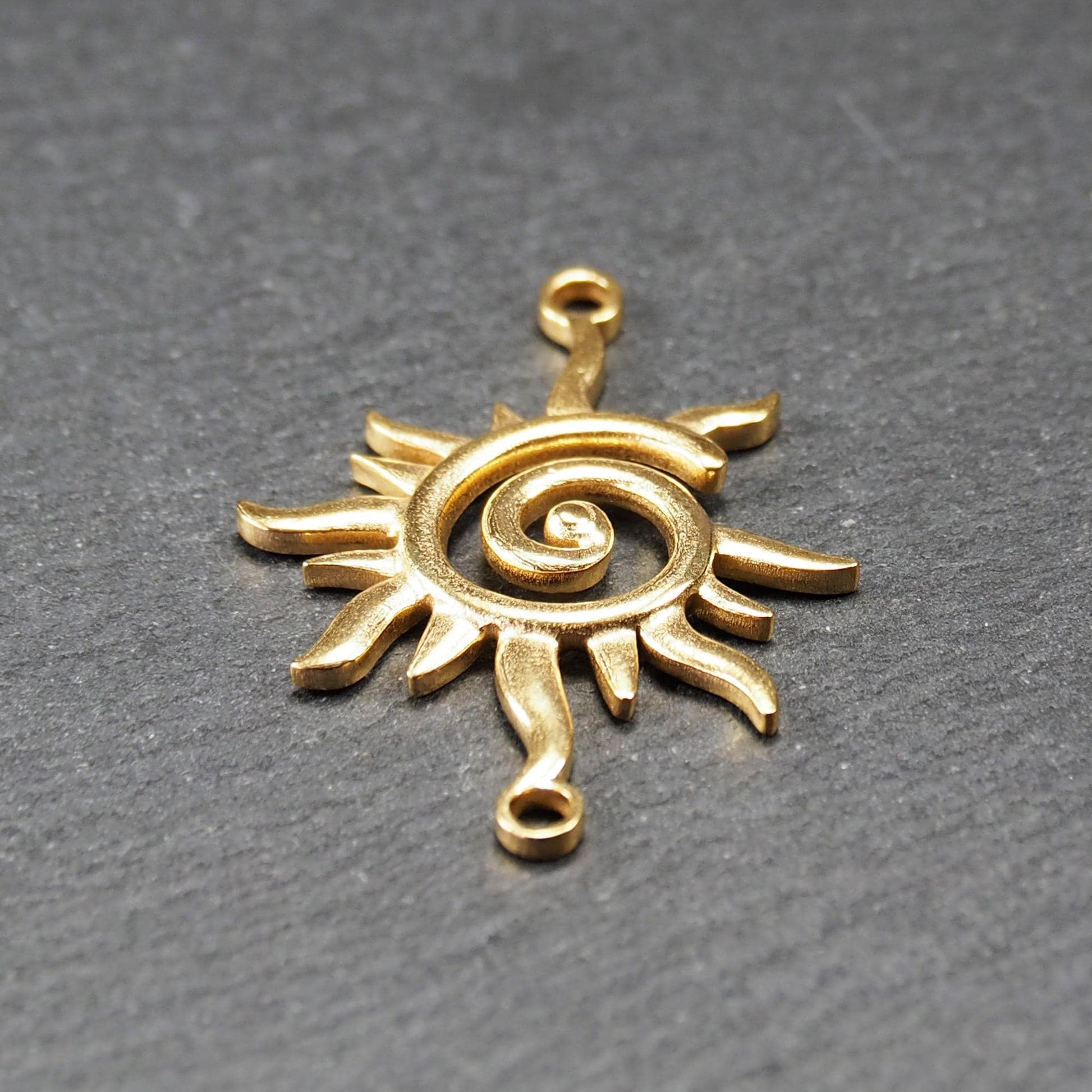 Goldfarbener, durchbrochener Sonnen-Verbinder mit spiralförmigem Zentrum, unregelmäßigen wellenförmigen Strahlen und je einer Öse oben und unten.