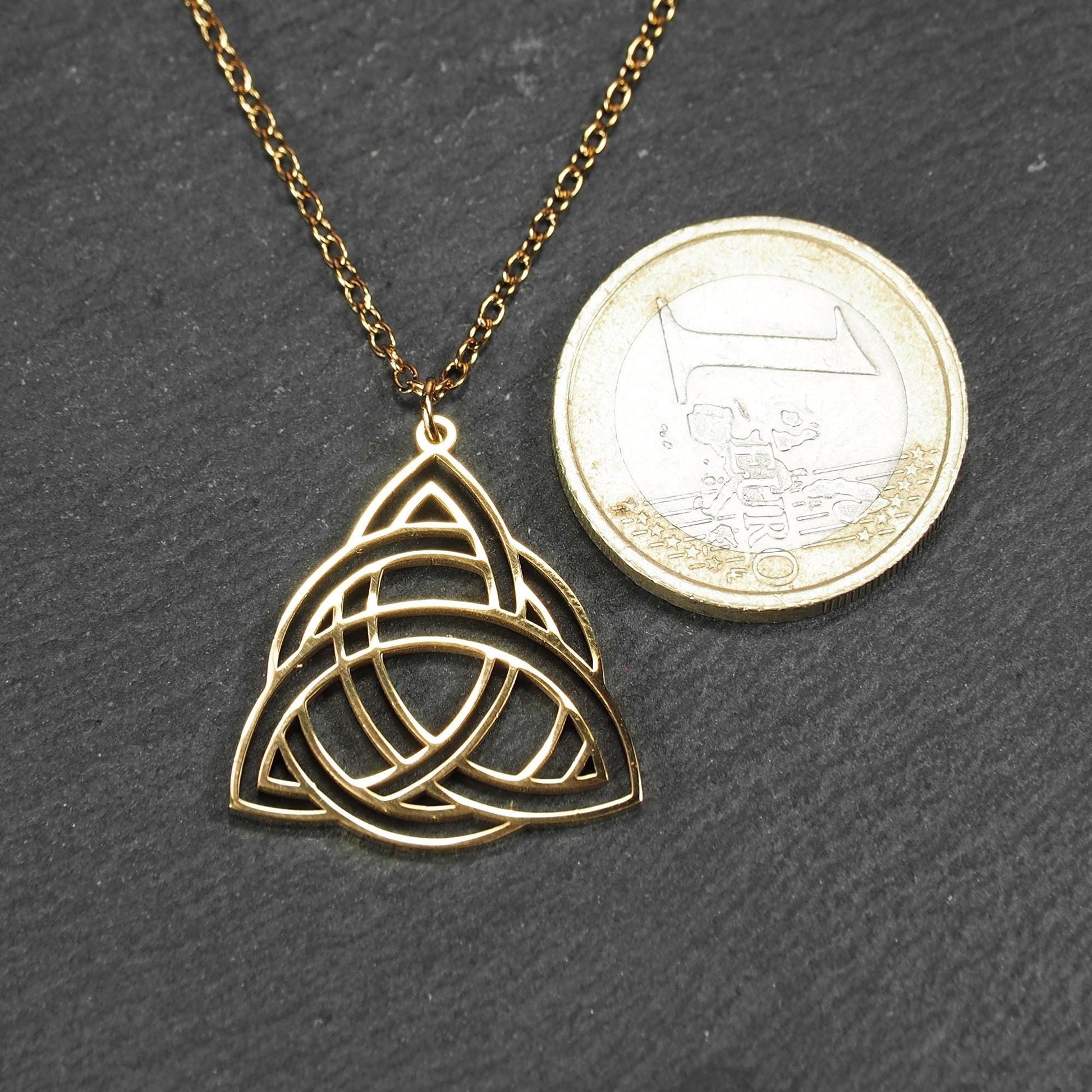 Vergoldeter Anhänger in offener Triquetra-Knotenform mit filigranen Durchbrüchen, an dünner goldfarbener Kette und sichtbarer Öse. 1-Euro-Münze als Größenvergleich.