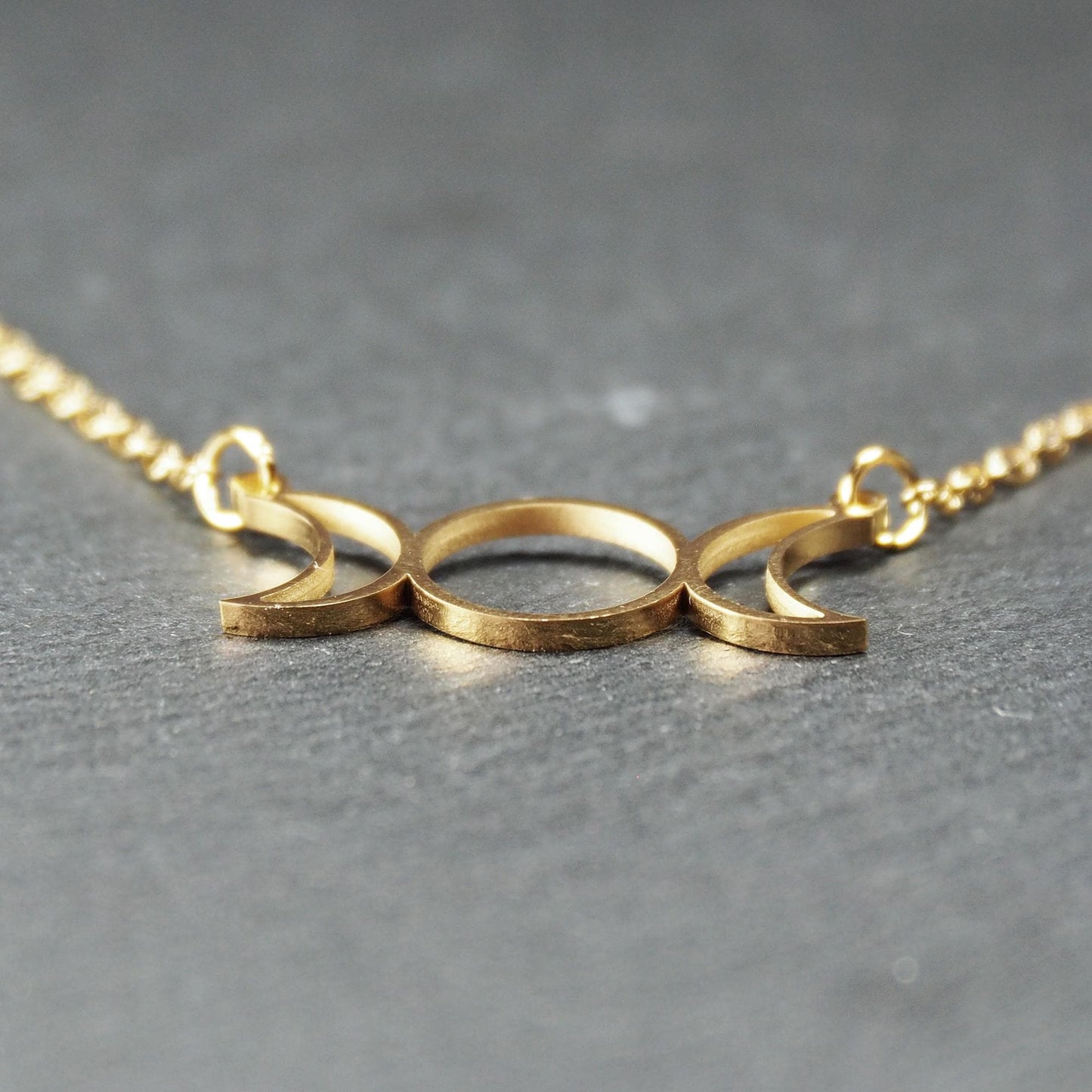 Collier mit goldfarbenem, offenem Anhänger in dreifacher Mondform (Mondsichel-Kreis-Mondsichel), an einer feinen Gliederkette mittels Ösen befestigt.