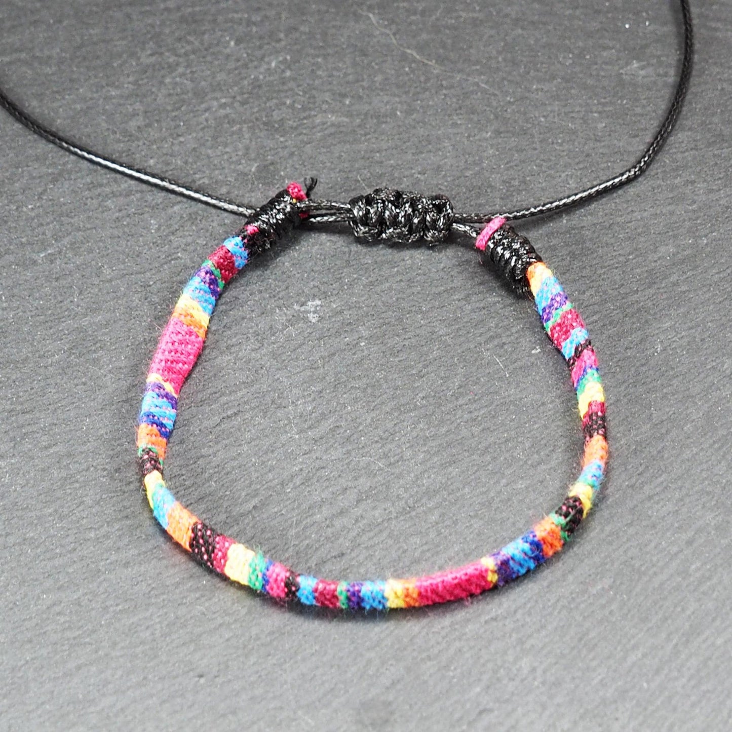 Gewebtes, flaches Armband mit regenbogenfarbenen Streifen, schmaler Form und feiner Webstruktur, schwarzer Schiebeknotenverschluss.